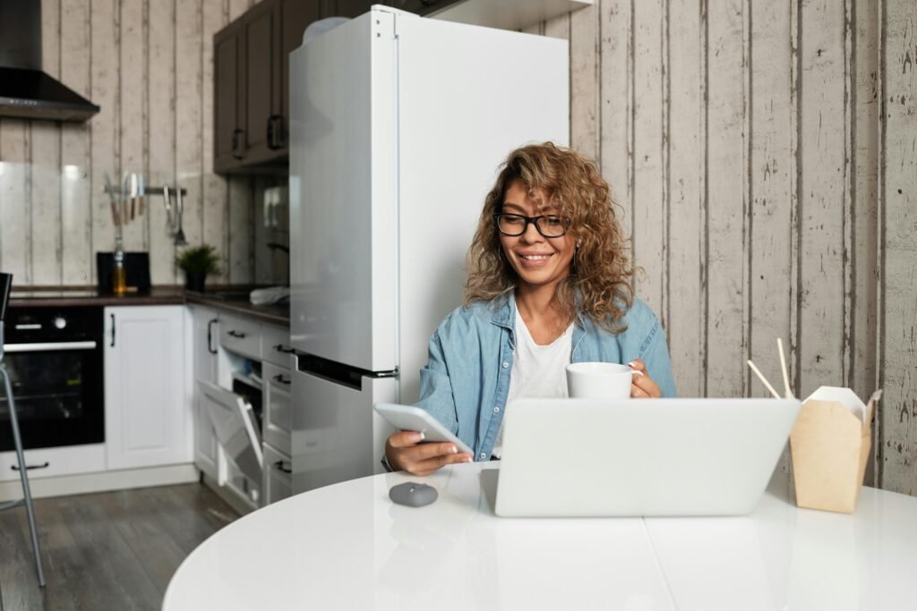 Woman using a smartphone and laptop while drinking coffee at a kitchen table.