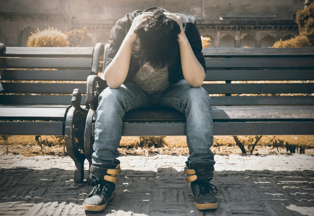 A young man experiencing depression sits on a park bench with his head in his hands, symbolizing emotional distress, fatigue, and the overwhelming nature of mental health struggles.